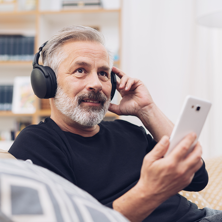 Mann mittleren Alters sitzt auf der Couch und hört mit dem Handy einen Podcast. | © iStock/stockfour