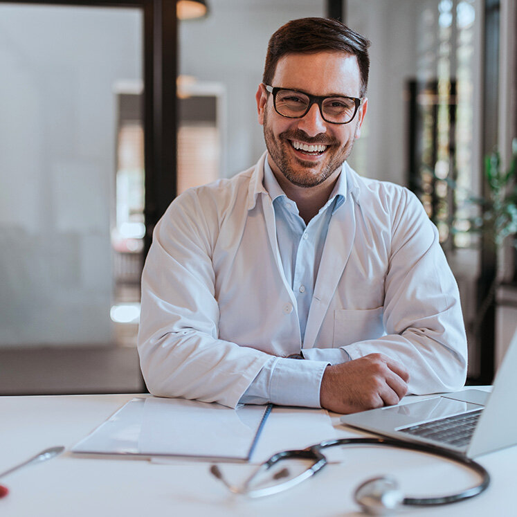 Jüngerer Hausarzt sitzt mit weißem Kittel an einem Schreibtisch vor seinem Laptop und lacht. | © iStock/nortonrsx