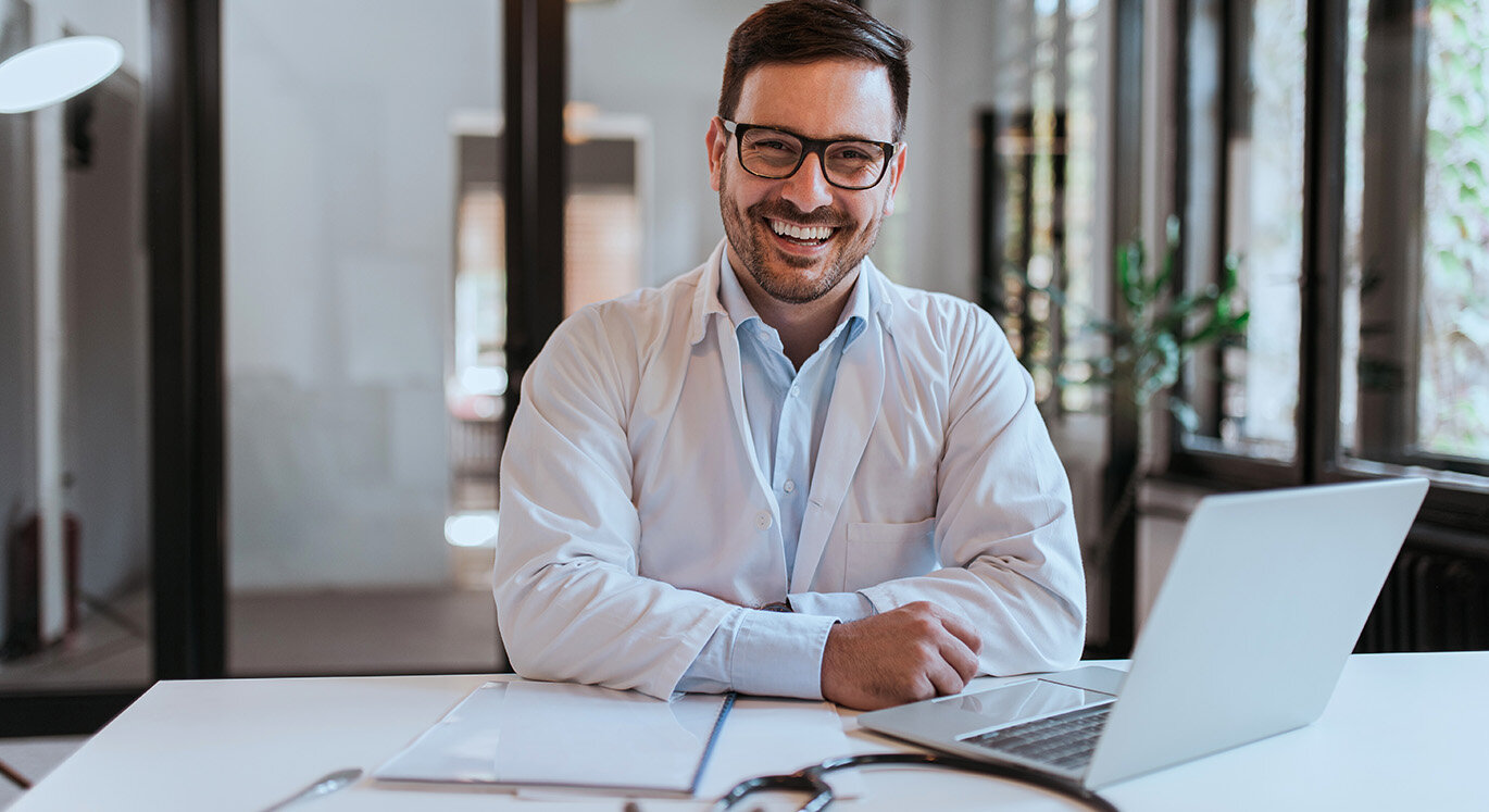 Jüngerer Hausarzt sitzt mit weißem Kittel an einem Schreibtisch vor seinem Laptop und lacht. | © iStock/nortonrsx