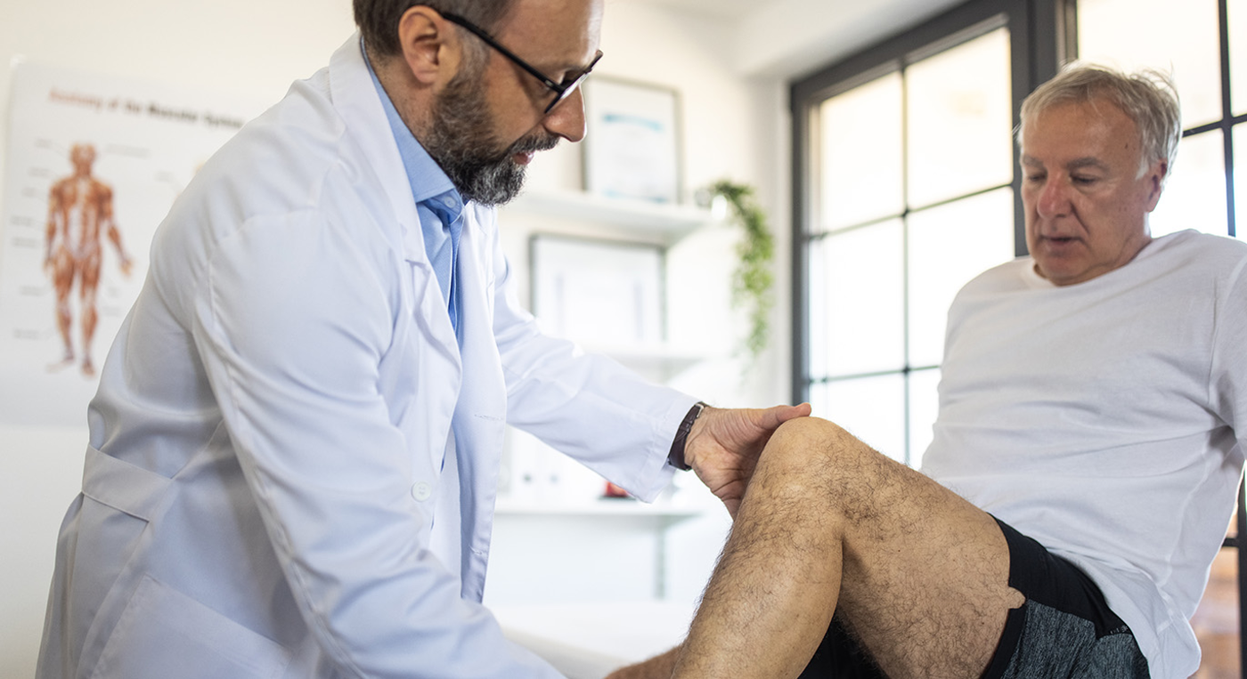 Hausarzt behandelt in seiner Praxis einen älteren Patienten mit Kniebeschwerden.