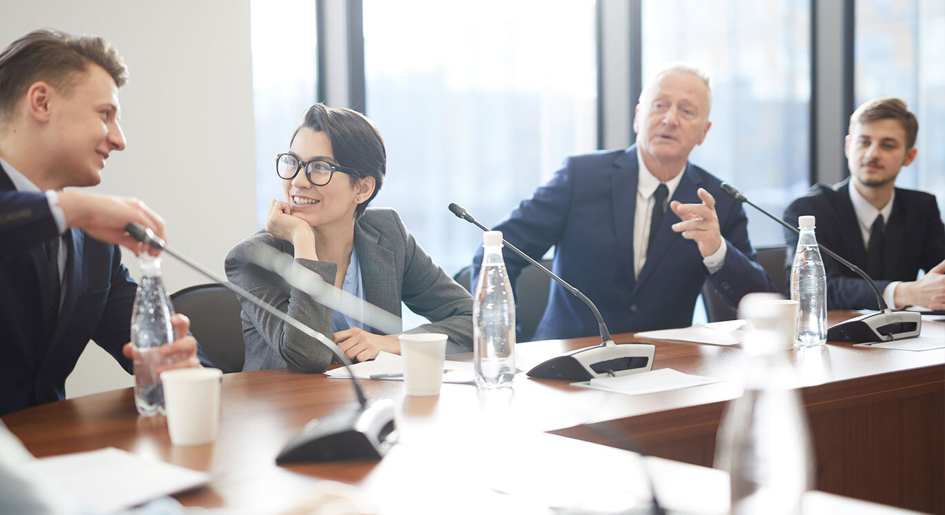 Drei Männer und eine Frau sitzen in einem Geschäftsmeeting an einem brauen Tisch. | © iStock/SeventyFour