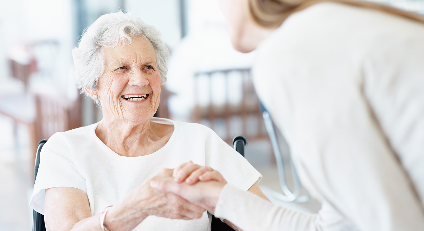 Ältere Patientin im Rollstuhl wird im Pflegeheim von ihrer Hausärztin begrüßt.