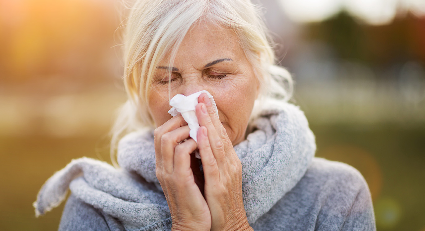 Ältere Patientin trägt einen Schal und putzt beim Spazierengehen im Herbst ihre Nase.