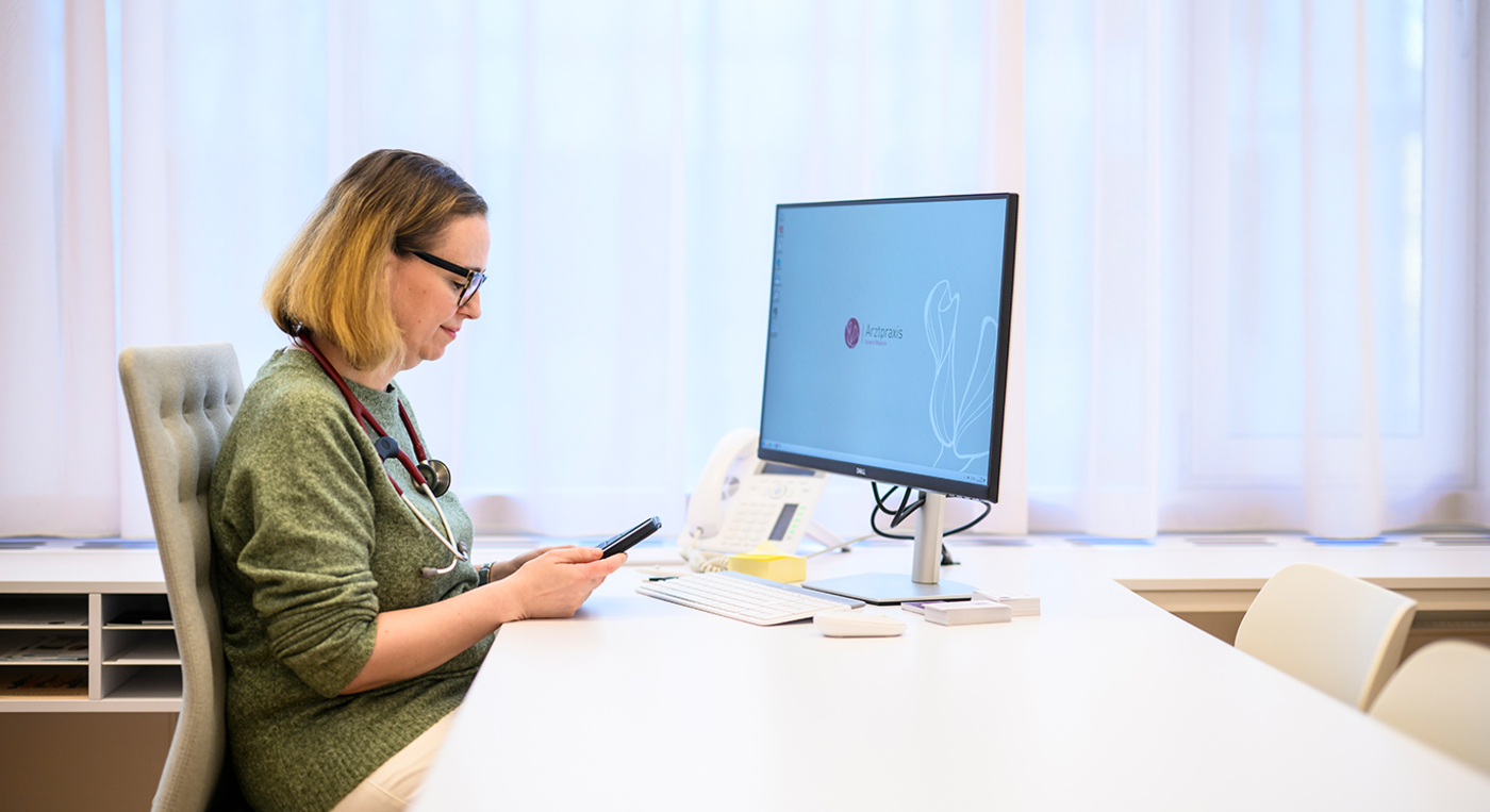 Hausärztin sitzt im Sprechzimmer und informiert sich am Smartphone.